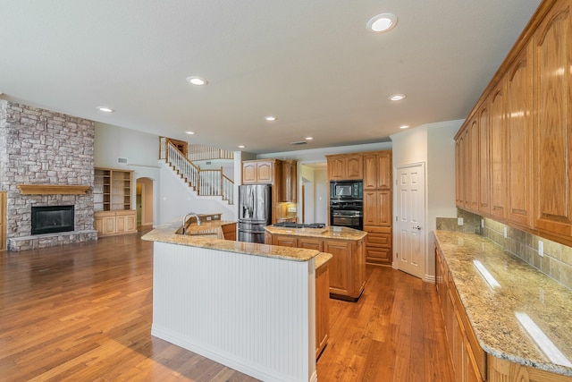 kitchen with black appliances, a stone fireplace, sink, a kitchen island with sink, and hardwood / wood-style flooring