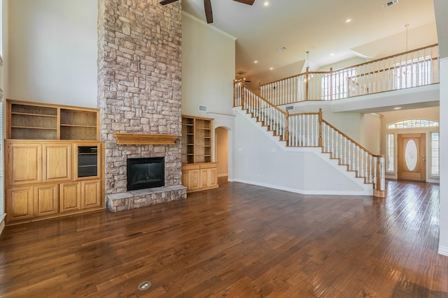 unfurnished living room with ceiling fan, a towering ceiling, a stone fireplace, and dark hardwood / wood-style floors