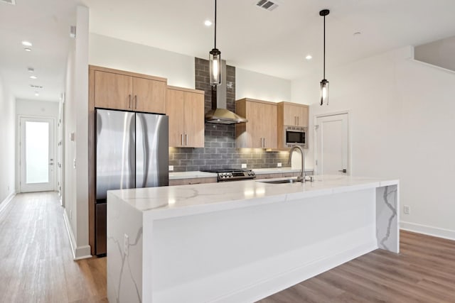 kitchen with an island with sink, a sink, decorative backsplash, appliances with stainless steel finishes, and wall chimney range hood