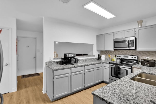 kitchen with stainless steel appliances, gray cabinetry, backsplash, light wood-type flooring, and light stone countertops