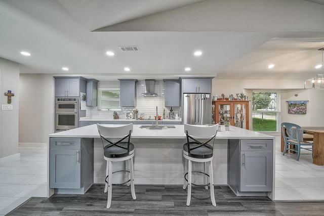 kitchen with wall chimney range hood, stainless steel appliances, tasteful backsplash, an island with sink, and a kitchen breakfast bar