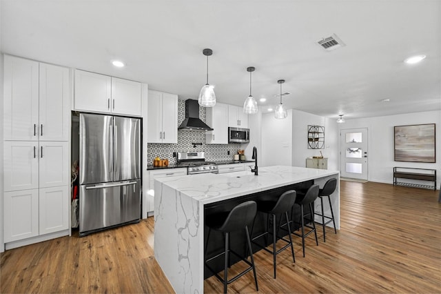 kitchen featuring appliances with stainless steel finishes, white cabinetry, a kitchen island with sink, light stone counters, and wall chimney exhaust hood