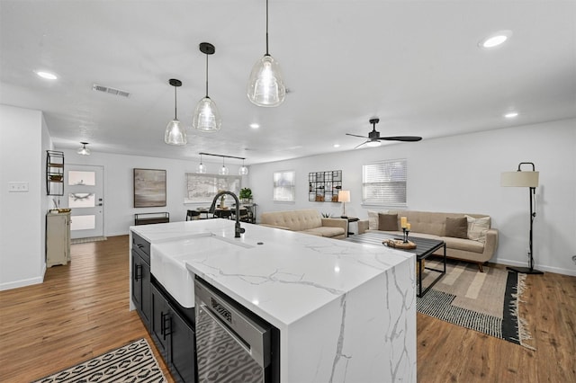 kitchen featuring sink, light stone counters, hanging light fixtures, dishwashing machine, and a kitchen island with sink