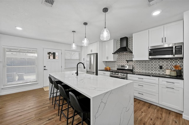 kitchen featuring white cabinets, appliances with stainless steel finishes, an island with sink, and wall chimney range hood