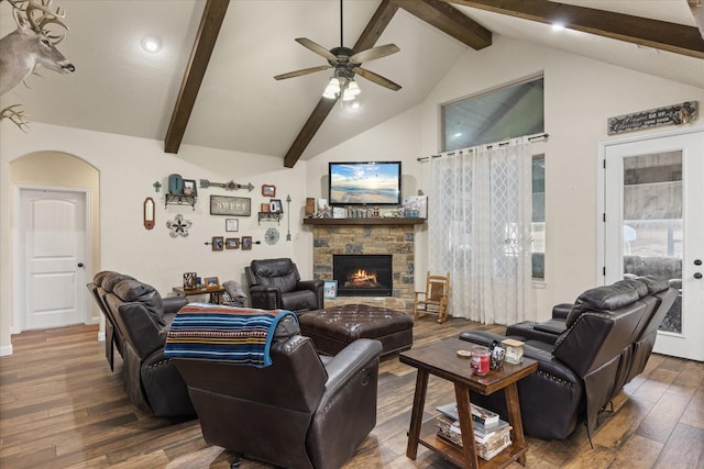 living room with ceiling fan, dark wood-type flooring, a stone fireplace, and vaulted ceiling with beams