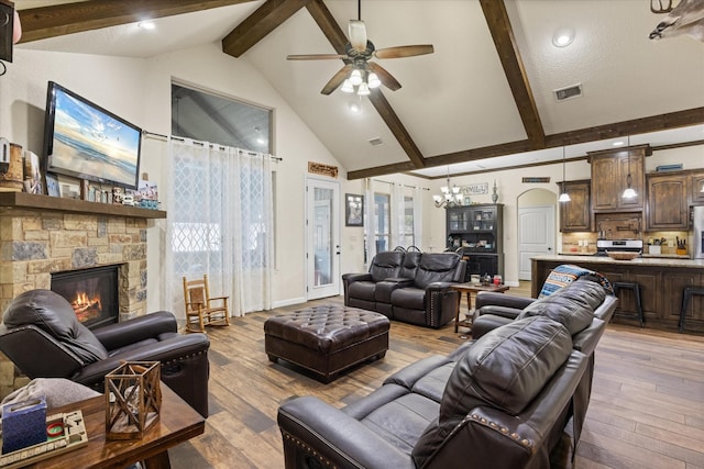 living room with beamed ceiling, light wood-type flooring, a stone fireplace, high vaulted ceiling, and ceiling fan with notable chandelier