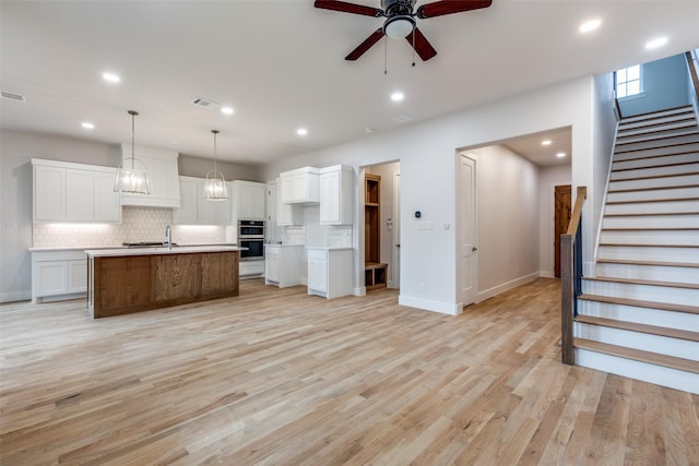 kitchen with white cabinets, decorative light fixtures, double oven, an island with sink, and ceiling fan