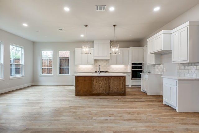 kitchen featuring white cabinetry, decorative light fixtures, a kitchen island with sink, double oven, and light hardwood / wood-style flooring