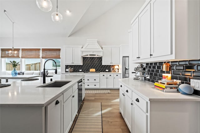 kitchen with vaulted ceiling, sink, hanging light fixtures, an island with sink, and custom range hood