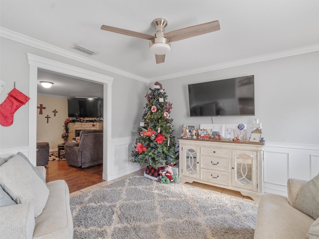 tiled living room with ceiling fan, a stone fireplace, and crown molding