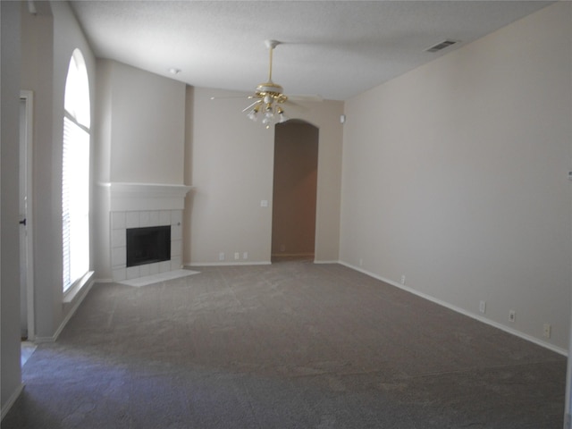 unfurnished living room with ceiling fan, a tiled fireplace, plenty of natural light, and dark colored carpet