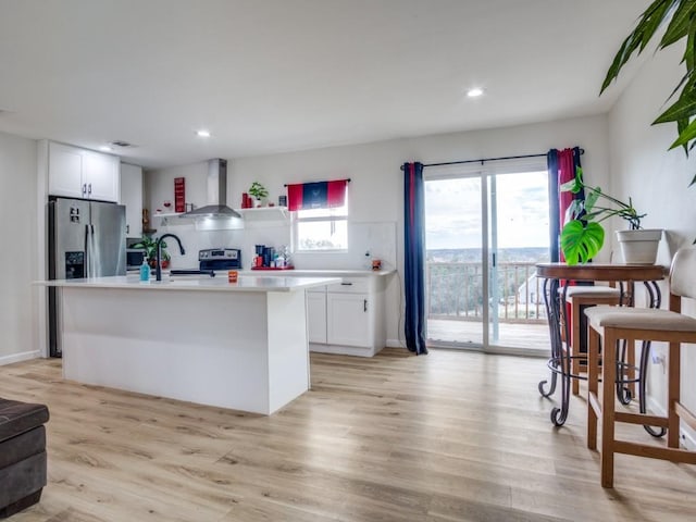 kitchen with white cabinets, wall chimney exhaust hood, an island with sink, and light hardwood / wood-style flooring