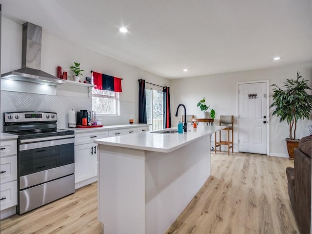 kitchen featuring wall chimney range hood, stainless steel electric range, sink, an island with sink, and white cabinets