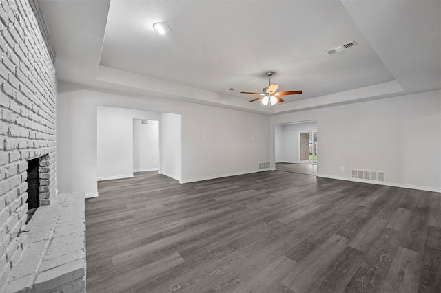 unfurnished living room featuring a brick fireplace, dark wood-type flooring, a tray ceiling, and ceiling fan