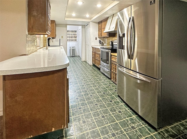 kitchen with appliances with stainless steel finishes, wall chimney exhaust hood, tasteful backsplash, sink, and a tray ceiling