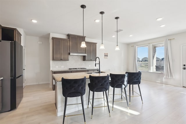 kitchen with sink, a kitchen island with sink, backsplash, stainless steel appliances, and decorative light fixtures