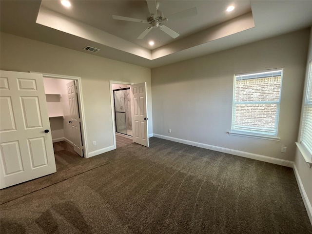 unfurnished bedroom with a raised ceiling, a walk in closet, ceiling fan, and dark carpet