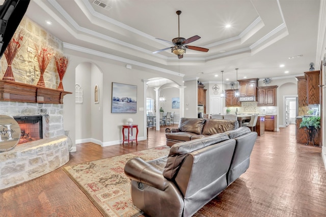 living room with a fireplace, ornamental molding, ceiling fan, a tray ceiling, and decorative columns