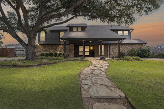 view of front facade featuring stone siding, a yard, a shingled roof, and fence