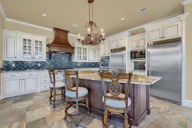 kitchen featuring wall chimney range hood, a center island with sink, light stone counters, and built in appliances