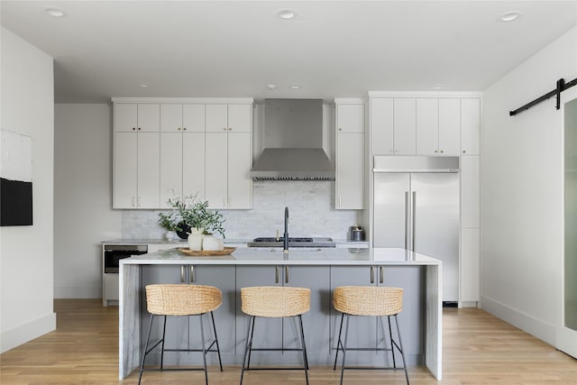 kitchen featuring a barn door, white cabinetry, an island with sink, built in refrigerator, and wall chimney exhaust hood