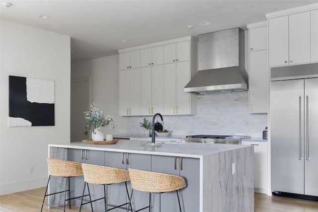 kitchen featuring white cabinetry, wall chimney exhaust hood, stainless steel built in fridge, and an island with sink