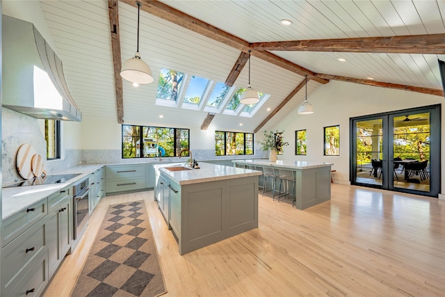 kitchen featuring a skylight, backsplash, a kitchen island with sink, wall chimney range hood, and pendant lighting