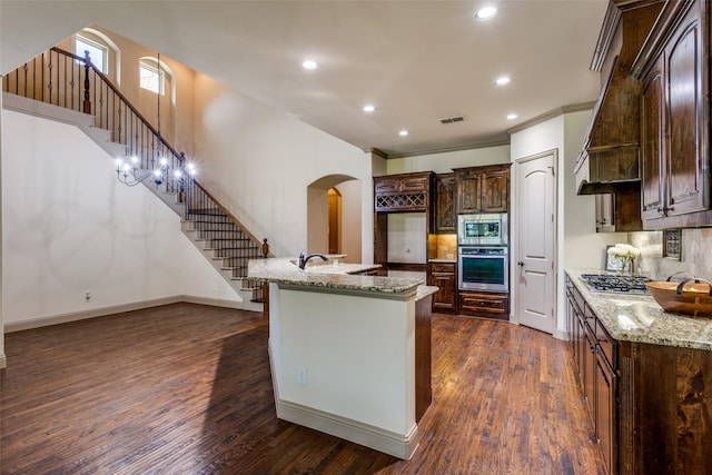 kitchen featuring an island with sink, stainless steel appliances, dark wood-type flooring, dark brown cabinets, and light stone countertops