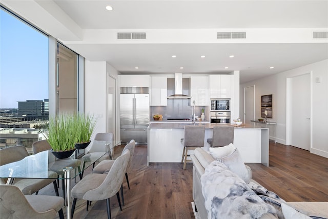 kitchen featuring an island with sink, dark hardwood / wood-style flooring, wall chimney range hood, white cabinets, and built in appliances