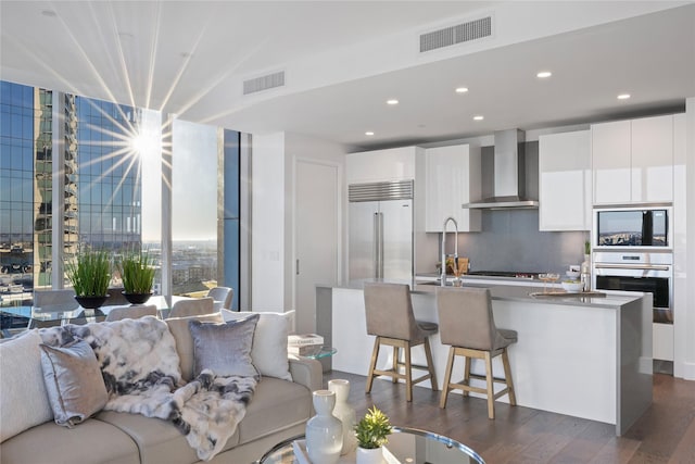 kitchen featuring wall chimney range hood, a center island with sink, built in appliances, white cabinetry, and dark wood-type flooring