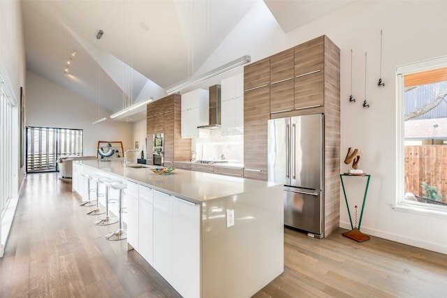 kitchen featuring white cabinets, wall chimney exhaust hood, stainless steel appliances, a center island with sink, and a breakfast bar area