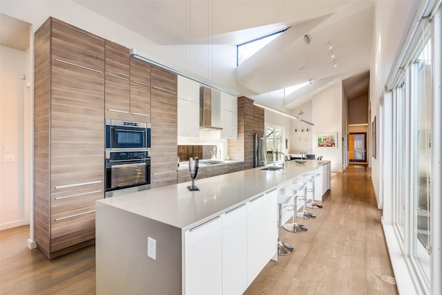 kitchen with a large island with sink, lofted ceiling, wall chimney exhaust hood, white cabinetry, and a kitchen breakfast bar