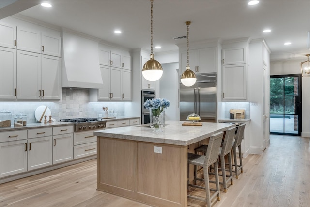 kitchen featuring white cabinetry, custom exhaust hood, appliances with stainless steel finishes, a kitchen island with sink, and hanging light fixtures