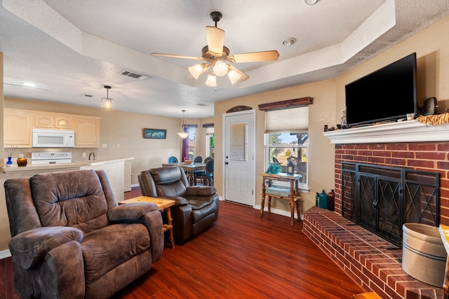 living room featuring a textured ceiling, dark wood-type flooring, ceiling fan, a brick fireplace, and a tray ceiling