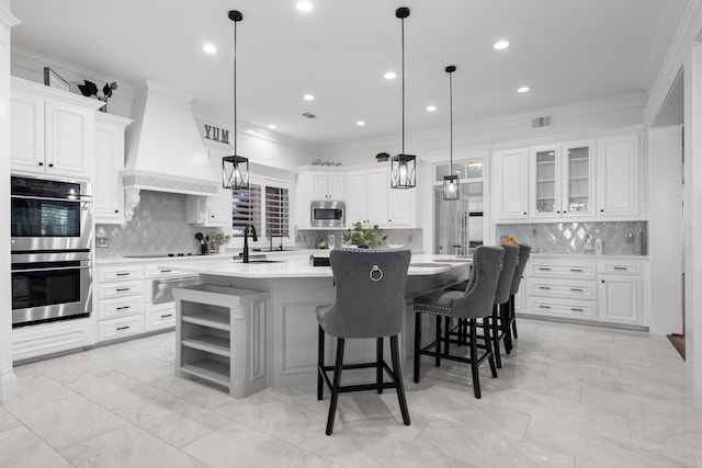 kitchen with premium range hood, stainless steel appliances, white cabinets, and a kitchen island with sink