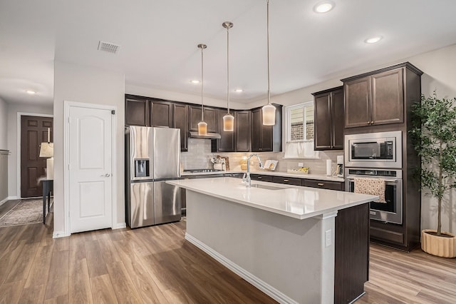 kitchen with pendant lighting, light hardwood / wood-style flooring, appliances with stainless steel finishes, an island with sink, and dark brown cabinets