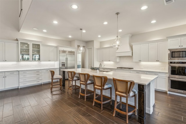 kitchen with premium range hood, a kitchen island with sink, built in appliances, pendant lighting, and a breakfast bar