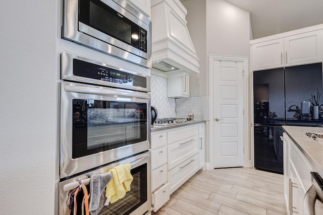 kitchen featuring premium range hood, stainless steel appliances, decorative backsplash, light wood-type flooring, and white cabinets