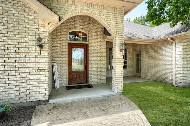 view of exterior entry with a patio area, brick siding, and roof with shingles