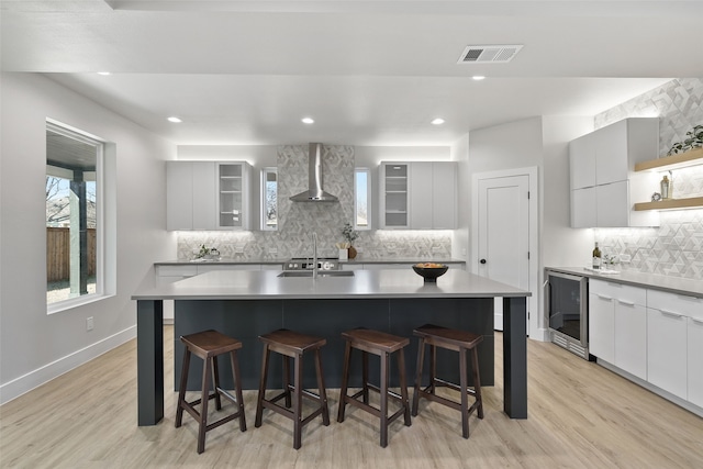 kitchen featuring an island with sink, a kitchen breakfast bar, beverage cooler, wall chimney exhaust hood, and light hardwood / wood-style flooring