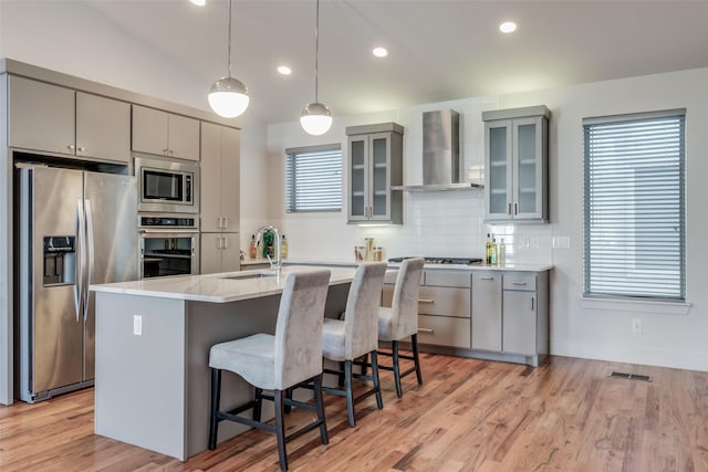 kitchen featuring wall chimney exhaust hood, decorative light fixtures, gray cabinetry, stainless steel appliances, and a center island with sink