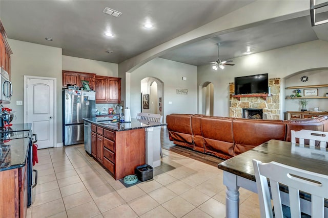 kitchen with ceiling fan, a fireplace, light tile patterned flooring, a kitchen island with sink, and stainless steel appliances