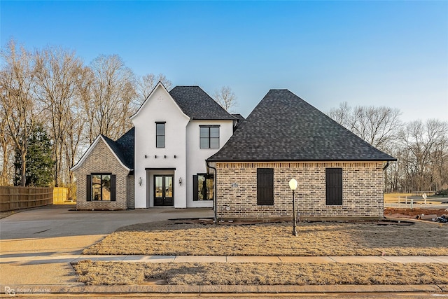 view of front facade with a shingled roof, stucco siding, fence, french doors, and brick siding