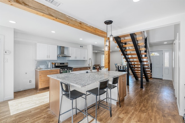 kitchen with white cabinetry, an island with sink, stainless steel gas range oven, and wall chimney range hood