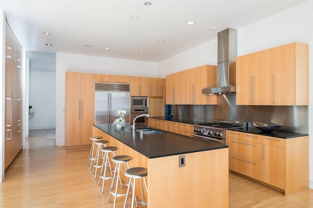 kitchen with tasteful backsplash, wall chimney range hood, a center island with sink, sink, and built in appliances