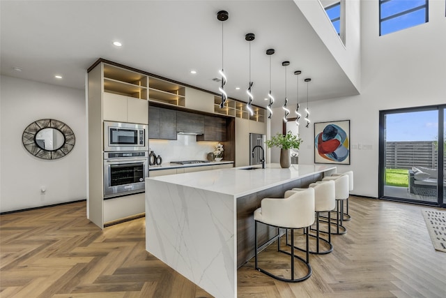 kitchen with an island with sink, modern cabinets, appliances with stainless steel finishes, hanging light fixtures, and light stone countertops