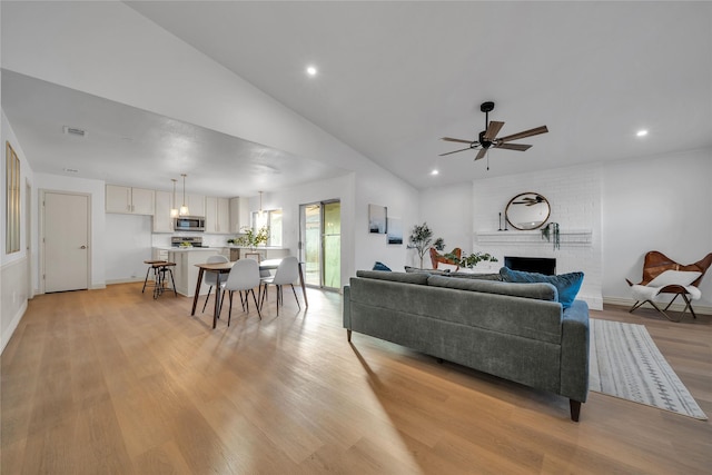 living room with ceiling fan, vaulted ceiling, a brick fireplace, and light hardwood / wood-style flooring