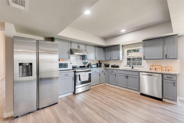 kitchen with sink, a tray ceiling, light wood-type flooring, light stone countertops, and appliances with stainless steel finishes