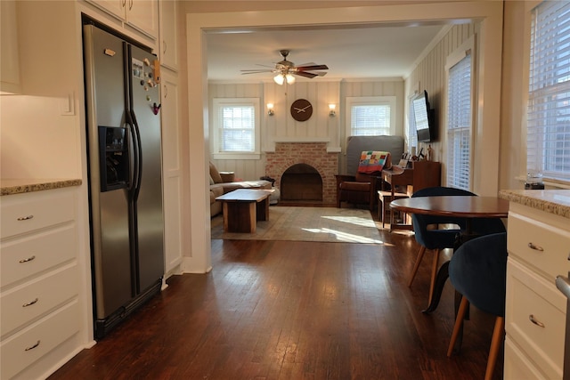 interior space with white cabinetry, wooden walls, ceiling fan, stainless steel fridge with ice dispenser, and a brick fireplace