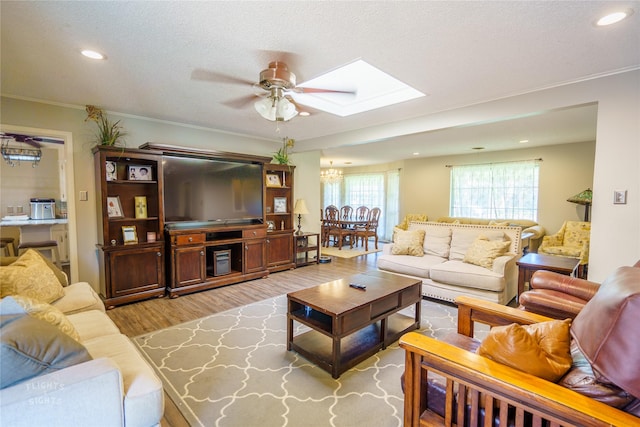 living room featuring ceiling fan with notable chandelier, a textured ceiling, a skylight, ornamental molding, and light hardwood / wood-style flooring